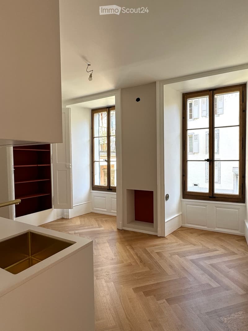 Empty room with light-colored wooden flooring, white walls, and large windows with wooden frames. A built-in shelf unit is visible on the left side.