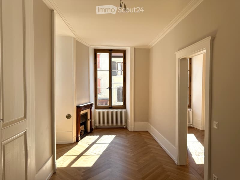 Empty hallway with wooden herringbone flooring, white walls, and a fireplace on the left side. Natural light streams in from a window at the end of the corridor.