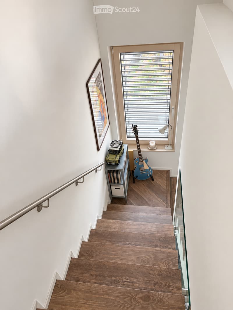 Wooden staircase with a metal handrail, white walls, a framed picture, a small shelf with books and a toy car, a blue electric guitar, and a window with blinds.