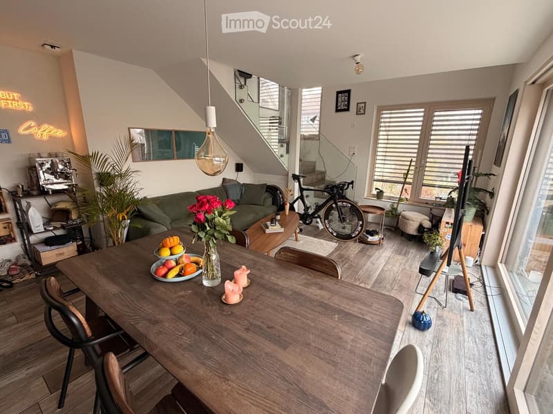 Bright living and dining area with wooden flooring, large windows, a green sofa, a wooden dining table with chairs, a bicycle, and decorative items including a vase with flowers and candles.