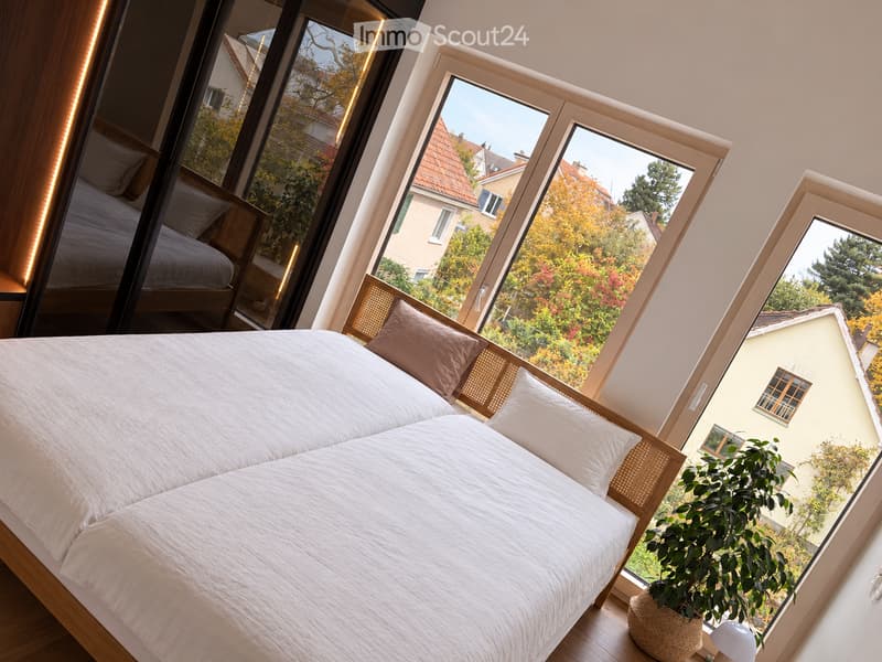 Modern bedroom with large windows offering a view of the garden and neighboring houses. Features a double bed with white bedding, a nightstand with a lamp, and a potted plant near the window.