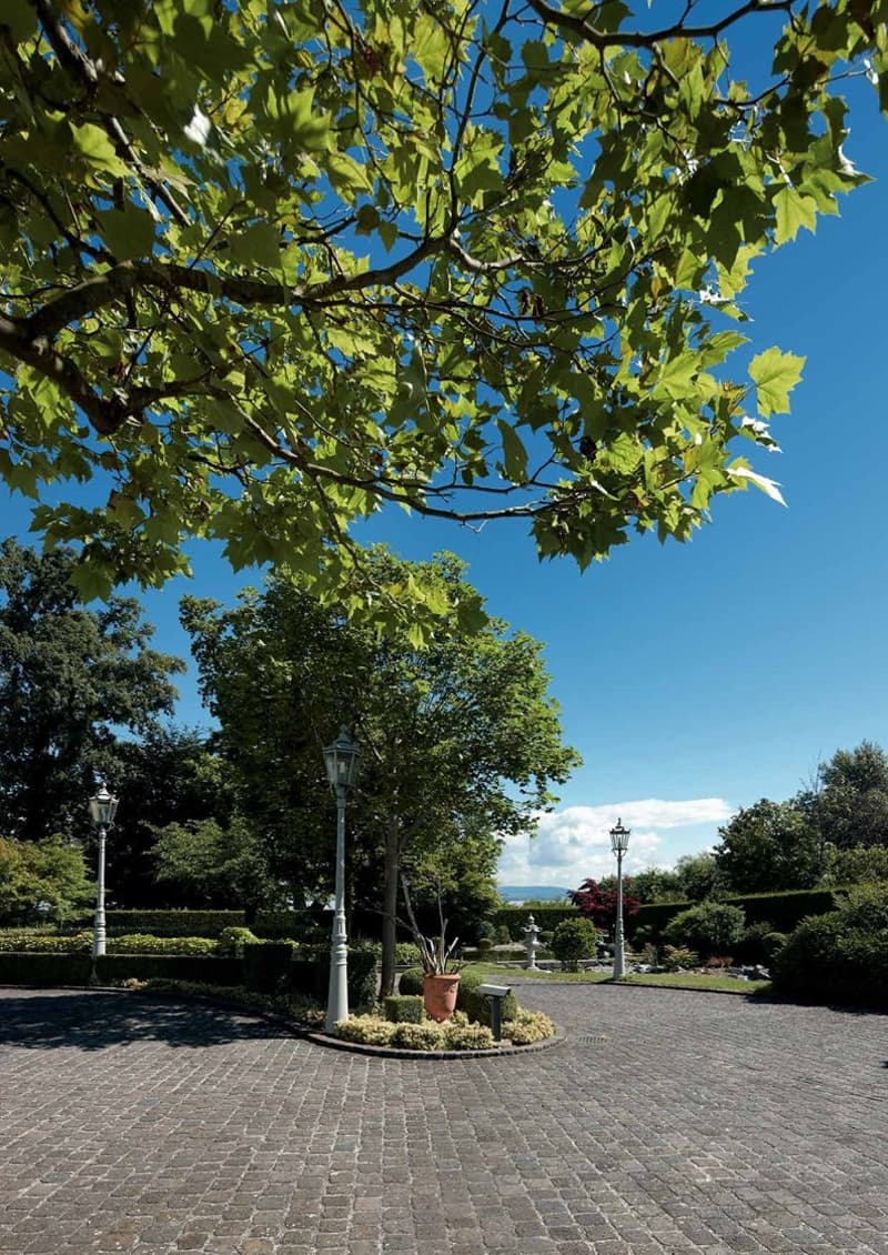 The image shows a well-maintained public garden or park with a cobblestone path, ornamental lamps, and a potted plant in the center. The garden is surrounded by lush green trees with vibrant leaves, creating a scenic and tranquil atmosphere. The backgroun