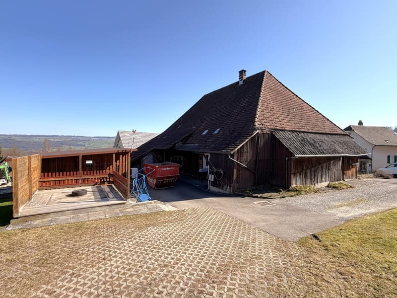 Traditional wooden barn with a red dumpster in the driveway, wooden fence, gravel driveway, and a view of the surrounding countryside.