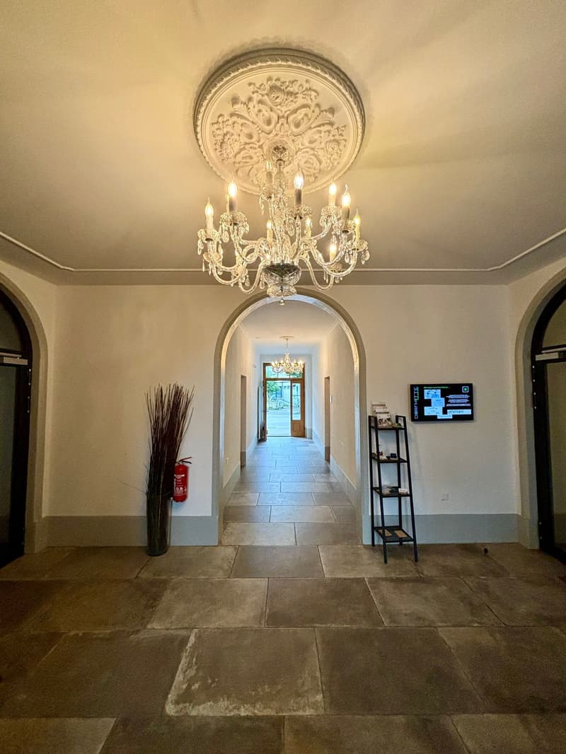 A hallway with a chandelier, tiled floor, arched doorways, a fire extinguisher, a TV, and a shelf with books