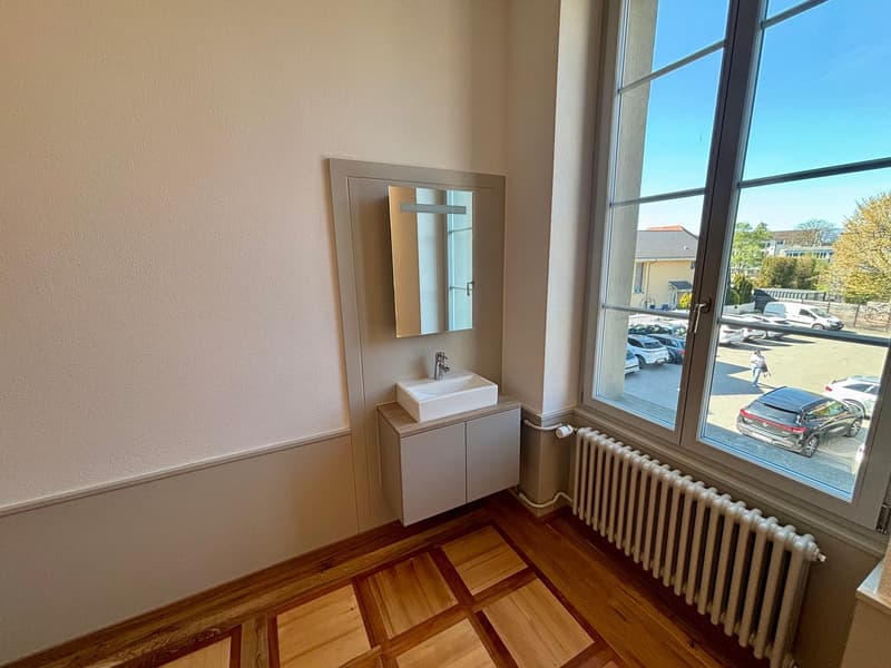 Bathroom with white sink, mirror, wooden floor, radiator, large window with view of parking lot