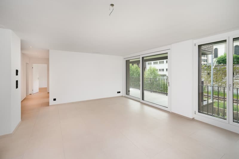 Spacious living room with large windows, sliding glass doors leading to a balcony, and a minimalist, open layout with white walls and light-colored flooring.
