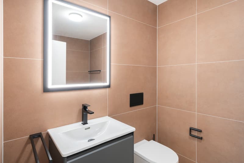 Modern bathroom with beige tile walls, a white sink vanity, a black faucet, and a large illuminated mirror.