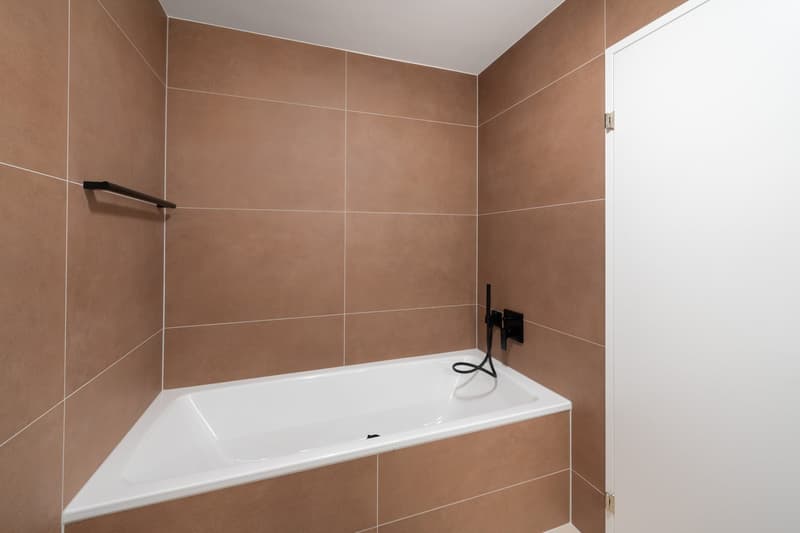 Spacious bathroom with brown tile walls, a white bathtub, and a black faucet fixture.