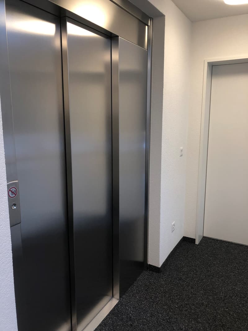 The image shows the interior of an elevator with two closed stainless steel doors. The floor is covered with a dark carpet. There is a no smoking sign visible on the wall.