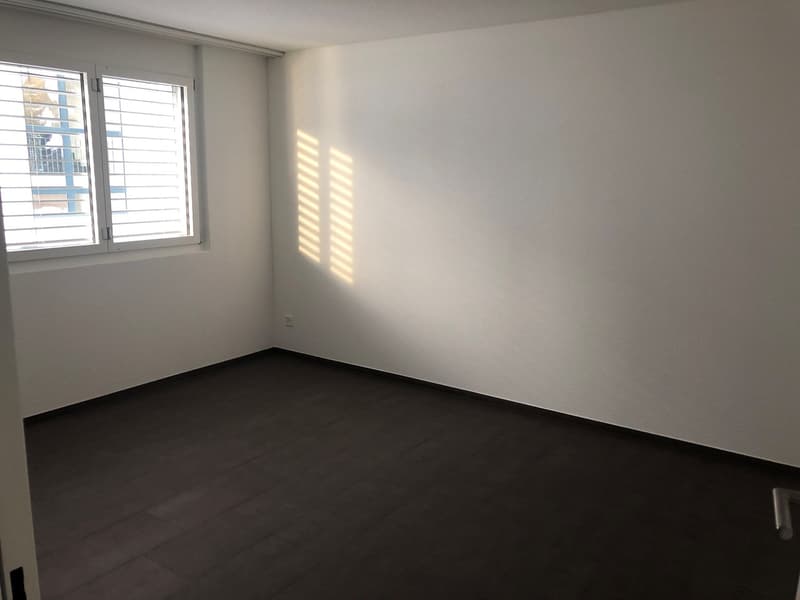 Empty bedroom with white walls, large window, and dark hardwood flooring.