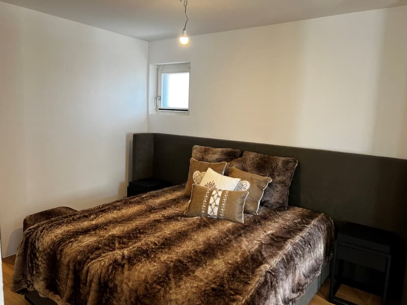 Spacious bedroom with a large bed covered in a plush, fur-like comforter and decorative pillows. The room has white walls and a small window.