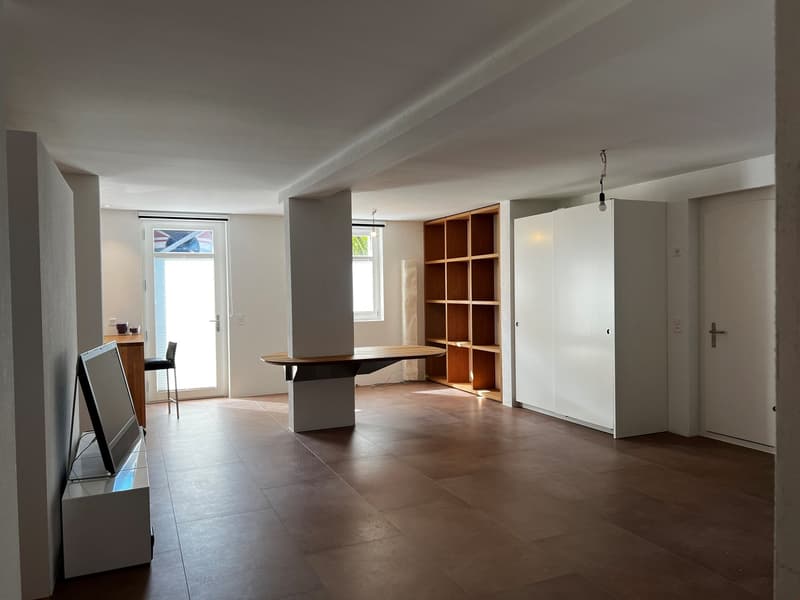 Spacious open-plan living area with hardwood flooring, white walls, large windows, built-in shelving unit, and a wooden dining table in the center of the room.