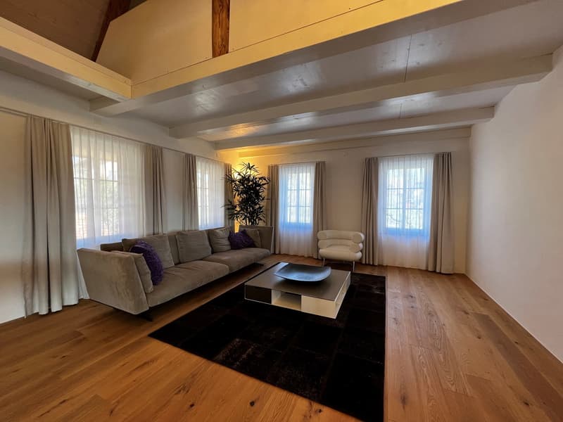 Spacious living room with high ceilings, exposed wooden beams, large windows, gray sofa, coffee table, and a potted plant. The room has a cozy and warm atmosphere.