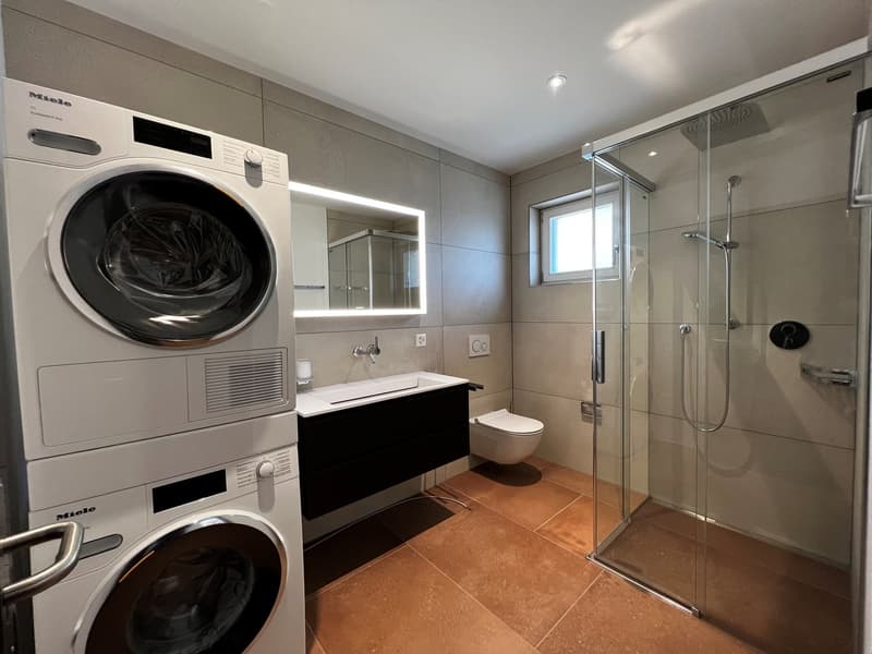 Spacious bathroom with a glass shower enclosure, a vanity with a sink, and two Miele washing machines stacked on top of each other. The room has a tiled floor and walls, providing a modern and clean look.