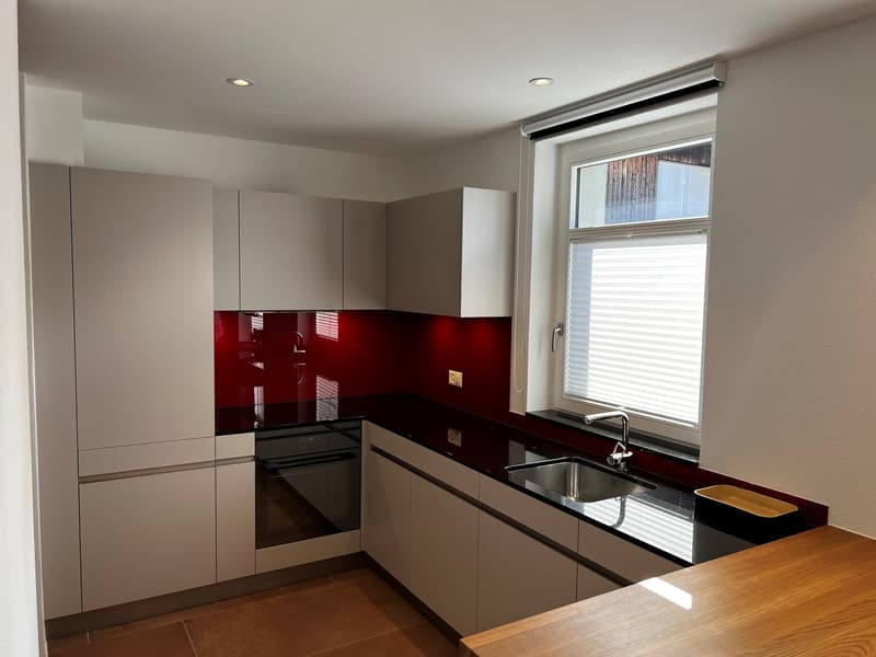 Modern kitchen with beige cabinets, red glass backsplash, black countertops, stainless steel appliances including a dishwasher, and a large window providing natural light.
