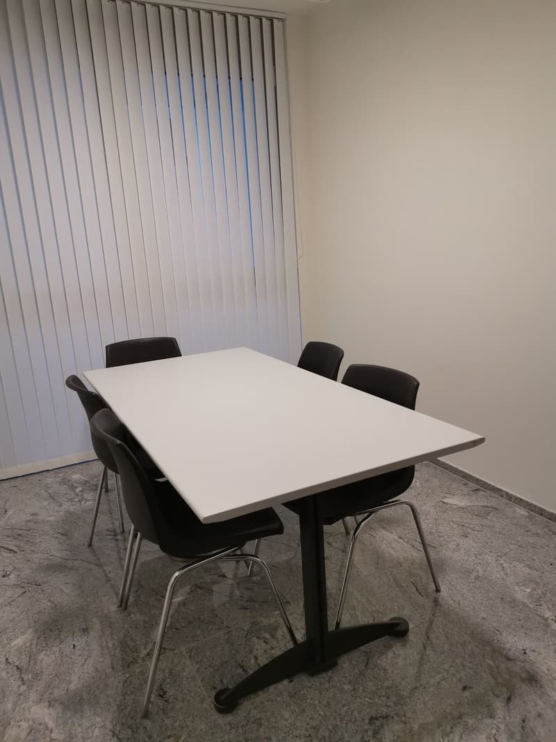 A room with white desk and black chairs, vertical blinds, and gray floor