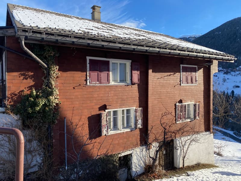 A three-story red brick house with a white wall, multiple shuttered windows, a brown roof with snow, a chimney, a small porch, and an outdoor space covered in snow with a mountain view.