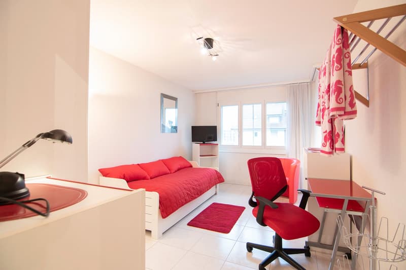 Small room with a red sofa bed, red rug, red chair, lamp, and desk. White walls and tiled floor.