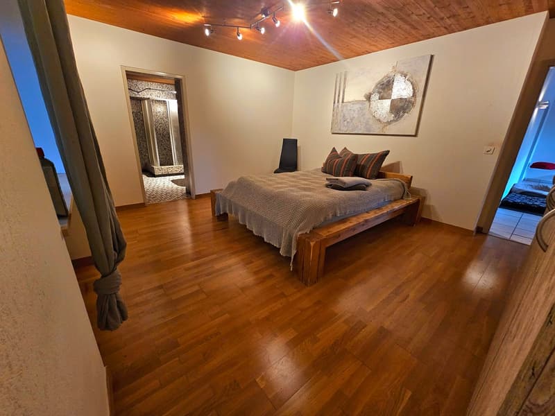 The image shows a cozy and well-decorated bedroom. The room has a wooden ceiling with track lighting, giving it a warm and inviting atmosphere. The focal point of the room is the large wooden bed frame with a plush comforter and decorative pillows. The ha