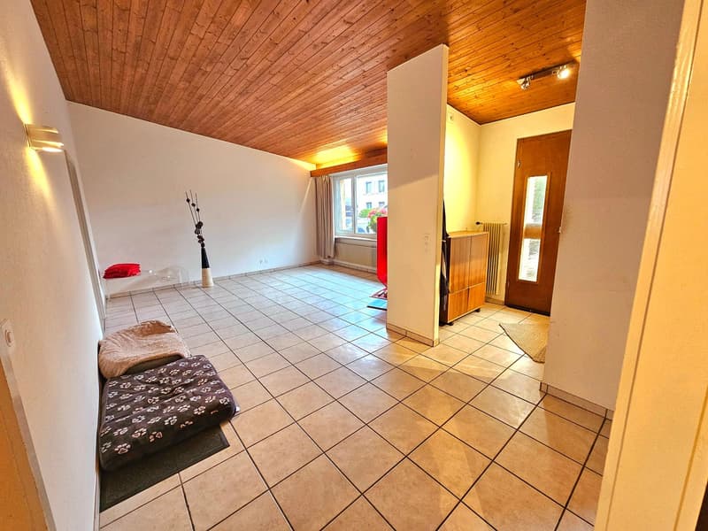 The image shows a spacious living room with a wooden ceiling and tile flooring. The room has a cozy and rustic feel, with a fireplace visible on the right side. The room is furnished with a few pieces, including a floral patterned cushion on the floor. Th
