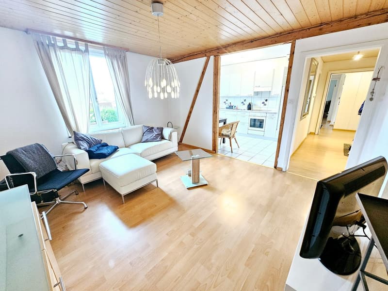 Spacious living room with hardwood floors, white walls, large windows, modern chandelier lighting, comfortable sofa and armchair, glass coffee table, and open access to the kitchen area.