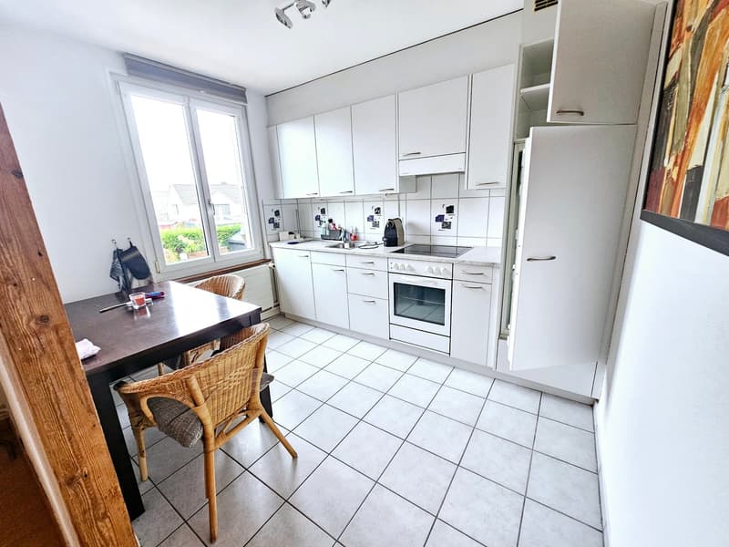 Spacious kitchen with white cabinets, granite countertops, built-in oven, and a dishwasher. The kitchen has a dining area with a wooden table and wicker chairs. Large windows provide natural light.