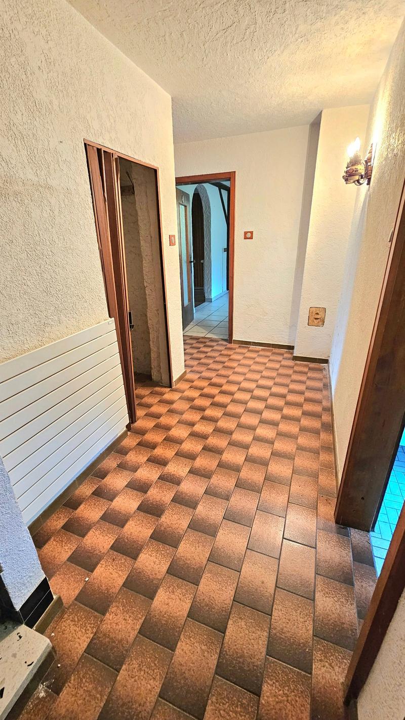 Corridor with brown checkered tiles, white walls, wooden doors, wall-mounted lamp. Two doorways leading to rooms, one with a wooden cabinet. Light shining through the hallway.