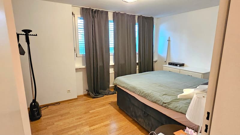 Bedroom with a large double bed, wooden floor, curtains, a nightstand with a lamp, an electric radiator, a table with books, and a floor lamp
