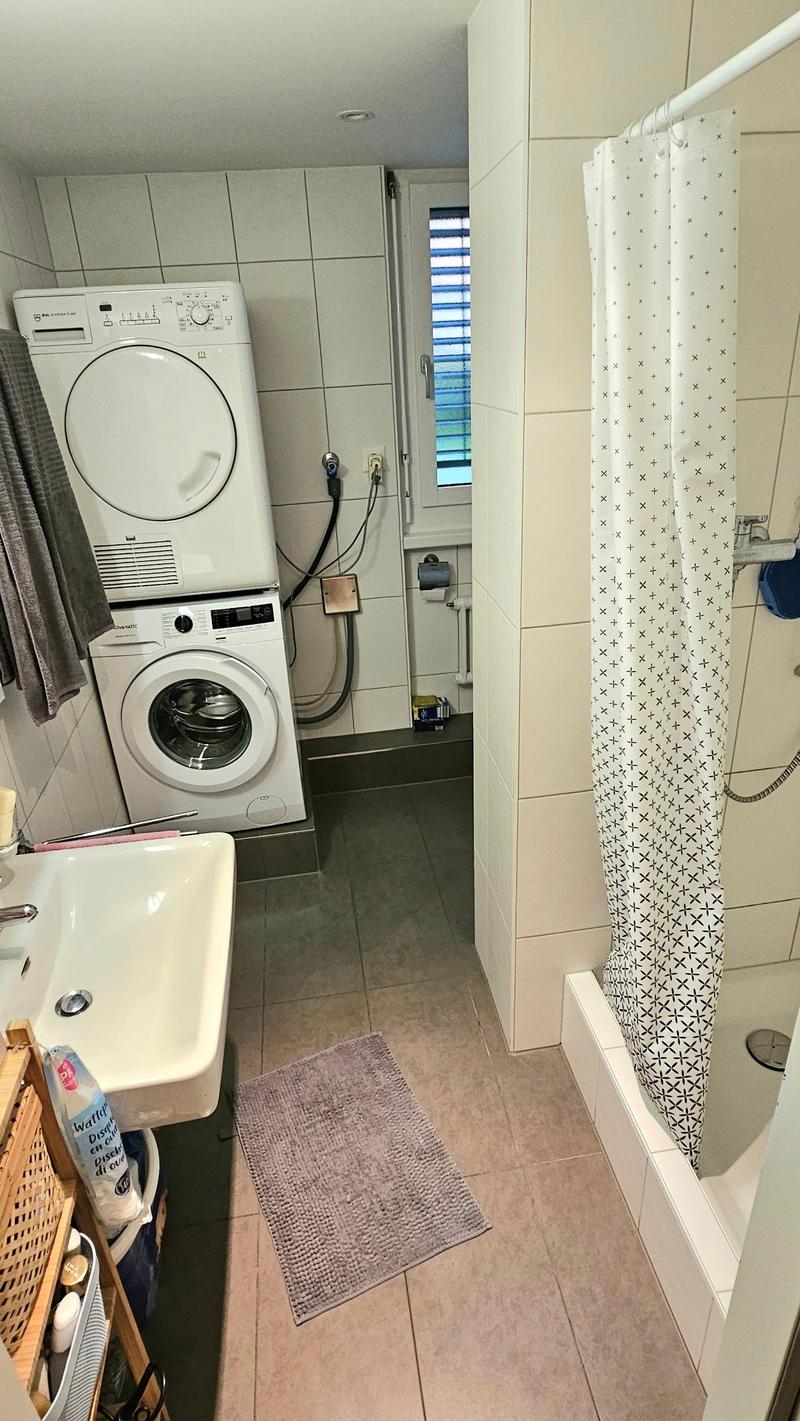 A small bathroom with white tiles, white sink, white bathtub, shower curtain, toilet roll holder, washing machine, and a door with a small window.
