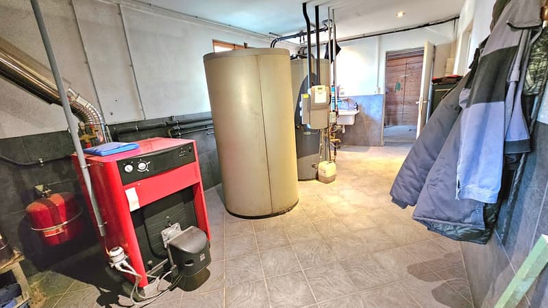 Utility room with boilers and pipes, red appliance with blue cover on top, red fire extinguisher, tiled floor, wooden door, sink, open door.