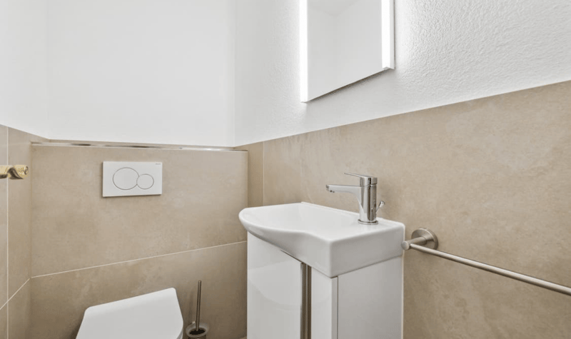 Clean, white modern bathroom with tile walls, white sink with chrome faucet, white toilet, small mirror mounted above sink.