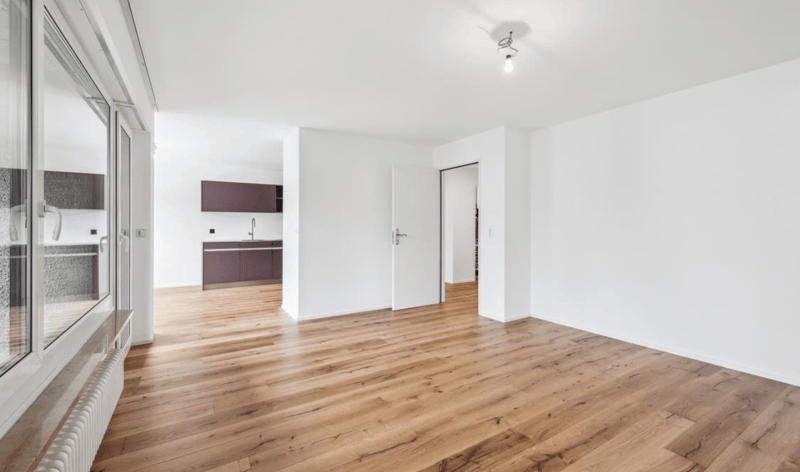 Empty room with hardwood floors, white walls, large glass doors, kitchen area, wooden cabinets, white ceiling