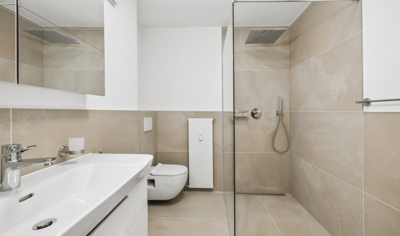 Modern bathroom with white tiled floors and walls, a glass-enclosed shower with a showerhead, a white vanity with a silver faucet, a white toilet, and a mirror