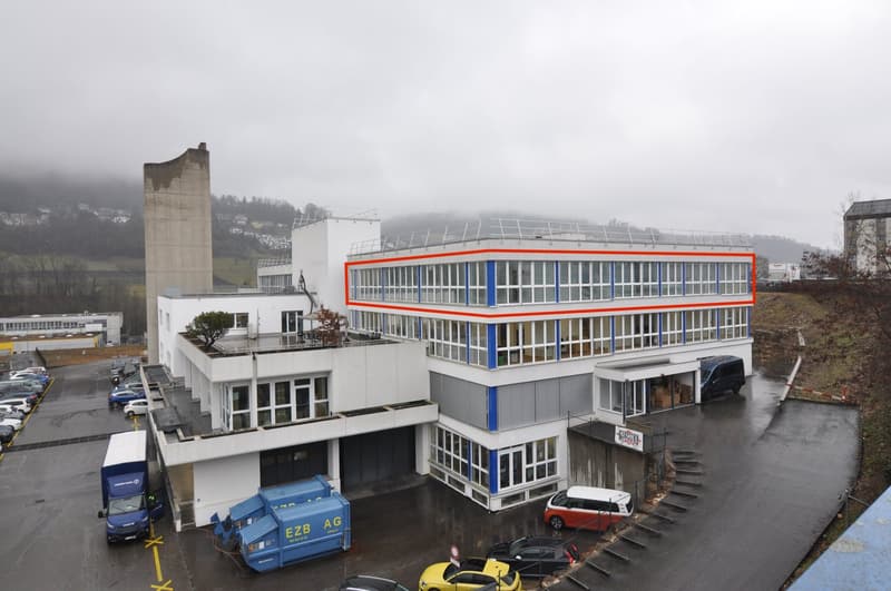Aerial view of a two-story building with a large parking lot in front. The building has glass windows and a red and blue outline highlighting a specific section. There is a blue truck parked near the building, with multiple cars parked in the lot. Behind