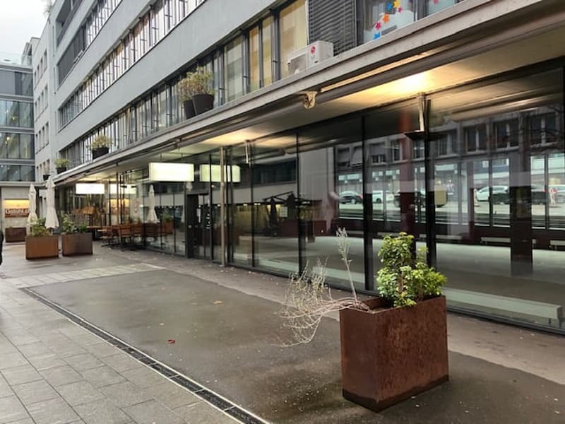 Modern building with large glass windows, balcony with plants, outdoor seating, central plant pot