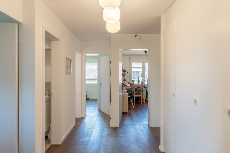 The image shows the interior of an apartment. The hallway has white walls and a hardwood floor. There are several doors leading to other rooms, including what appears to be a kitchen or living area visible through one of the open doorways. The hallway is
