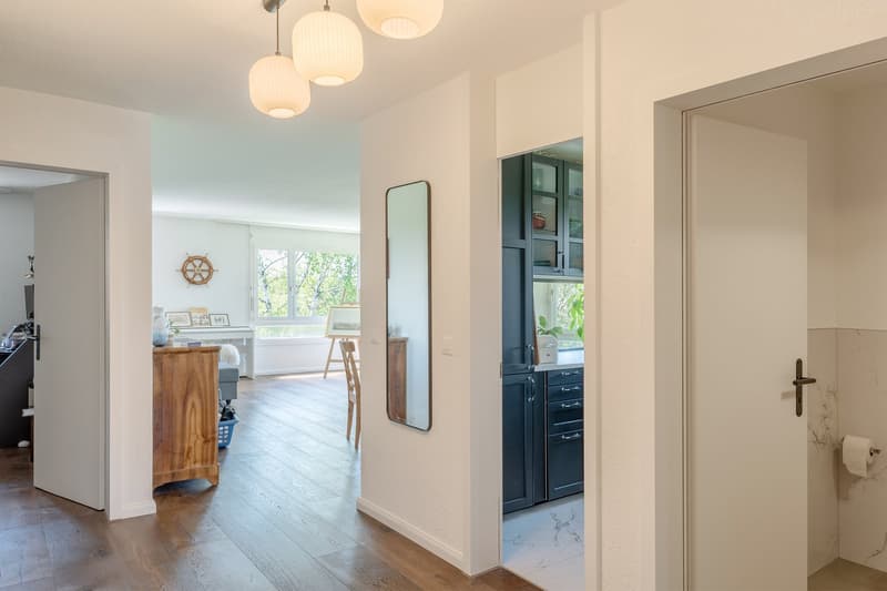 Spacious living room with hardwood floors, white walls, large windows, and a balcony. The room features a wooden cabinet, a mirror, and pendant lighting fixtures.