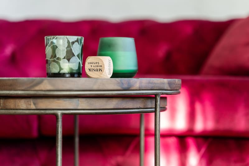 A wooden table with metal legs, topped with a small wooden block, and two decorative candles of different colors and patterns. Behind the table is a red velvet couch with pillows.