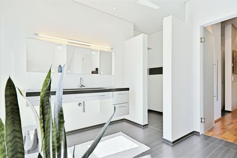 modern bathroom with double sinks, mirrors, cabinets, and a toilet, gray floor, white walls, open door to hallway