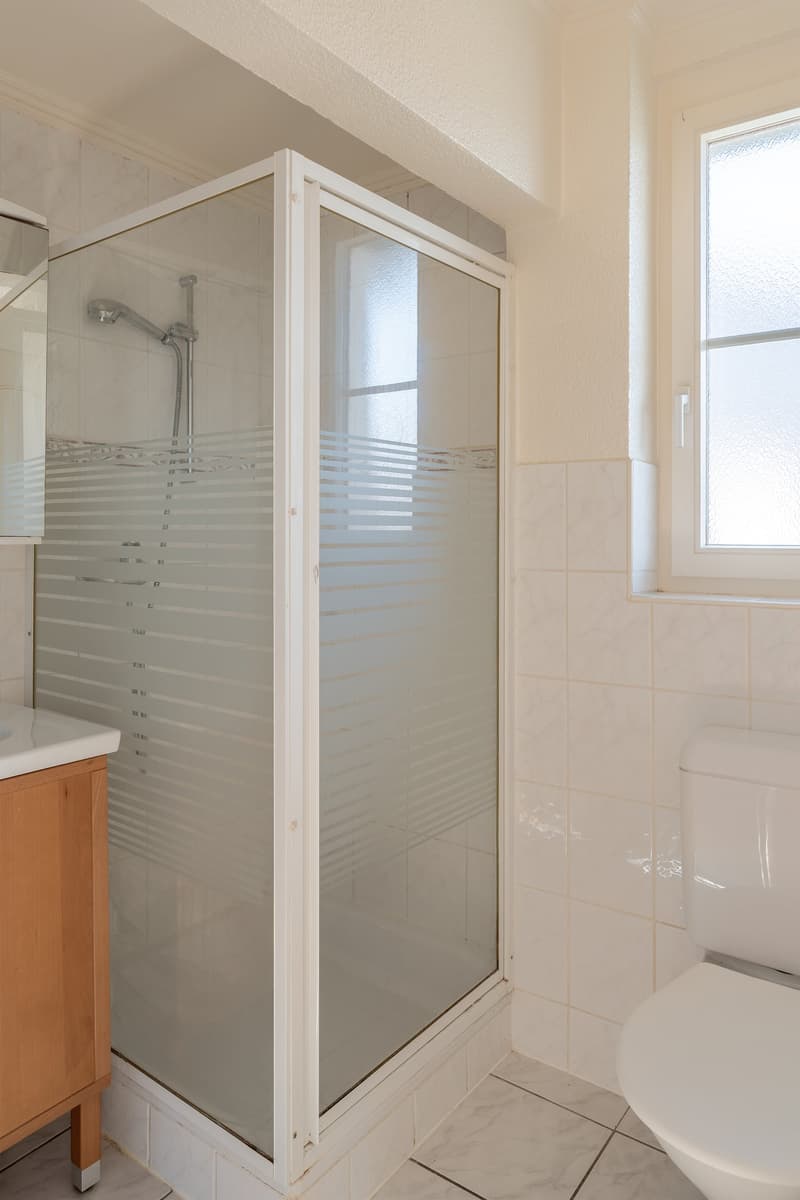 white tiled walls, white tiled floor, glass shower enclosure, white toilet, wooden cabinet, white sink