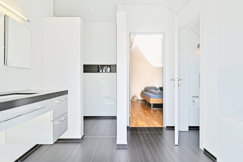 Minimalist bathroom with dark tiles, white walls, modern vanity, and an open doorway leading to a bedroom.