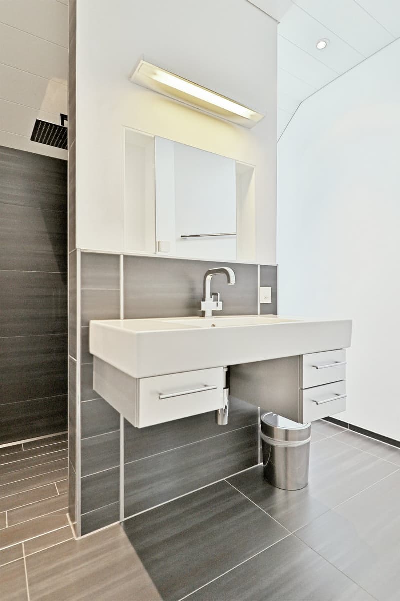 modern bathroom with white sink, grey tiled floor, built-in mirror, shower, and trash can
