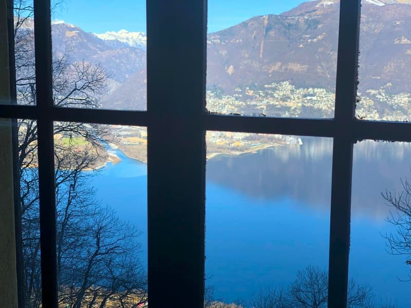 The image shows a scenic view of a lake surrounded by mountains, as seen through a window frame. The lake appears to be a deep blue color, reflecting the sky and the surrounding landscape. The mountains in the background are snow-capped, indicating a moun