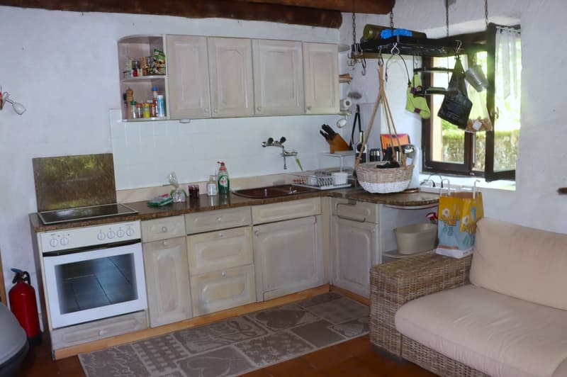The image shows a rustic kitchen with white cabinets, a gas stove, a sink, and various kitchen utensils and appliances. There is a wicker basket and a sofa visible in the corner of the room.
