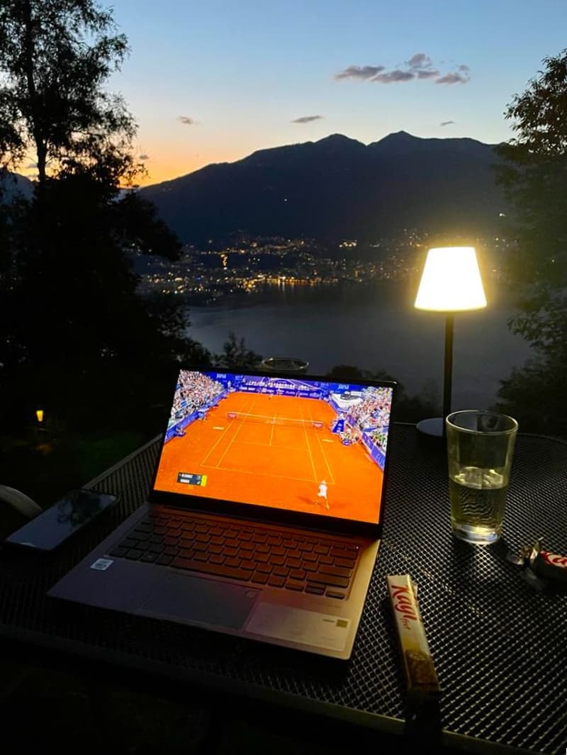 The image shows a laptop displaying a tennis match on a court, set against the backdrop of a scenic mountain landscape at sunset. A glass of drink and a snack are also visible on the table, suggesting a relaxing outdoor setting to enjoy the match.