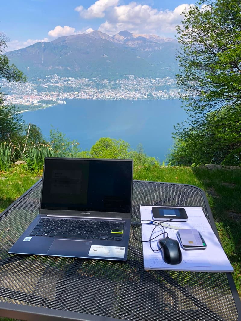 Laptop, mouse, and phone set up on a table with a scenic view of a lake surrounded by mountains and trees in the background.