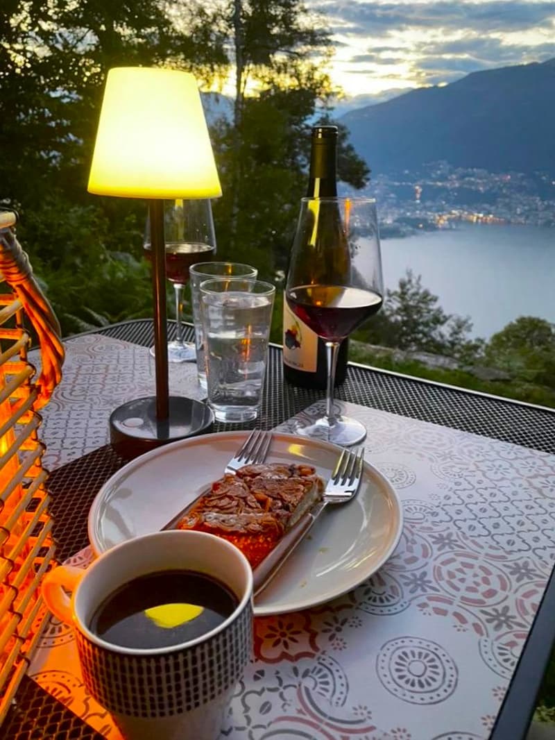 Outdoor dining table with a plate of food, wine glasses, and a bottle of wine. The table is set against a backdrop of a scenic lake or body of water surrounded by mountains and trees. A lamp provides lighting for the evening setting.