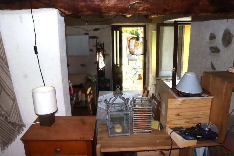 The image shows a cozy and rustic interior space, with a wooden ceiling and exposed beams. There is a small refrigerator, a table with various items on it, and a lamp. The room appears to have a door leading to an outdoor area.