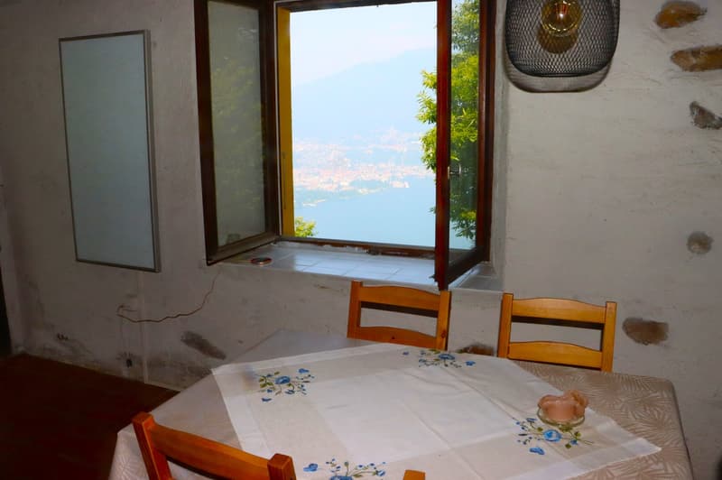 This image shows a room with a large window overlooking a scenic view. The room has a table with a floral tablecloth and two wooden chairs. There is also a mirror on the wall and a decorative light fixture. The overall atmosphere appears to be cozy and in