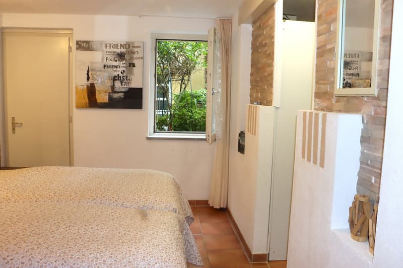 Bedroom with double bed, window, white walls, brick wall, radiator, painting, mirror, open door
